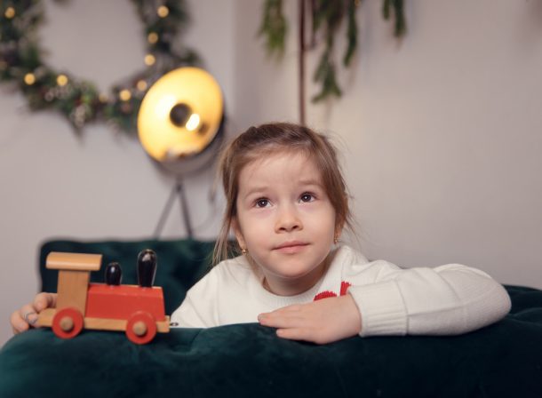 Fetiță visătoare într-un pulover alb, rezemată de o canapea din catifea verde și ținând un trenuleț roșu de jucărie, într-un decor de Crăciun