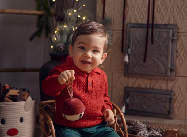 Băiețel vesel pe un scăunel din ratan, arătând un glob festiv, într-un decor cald de Crăciun lângă o sobă din teracotă
